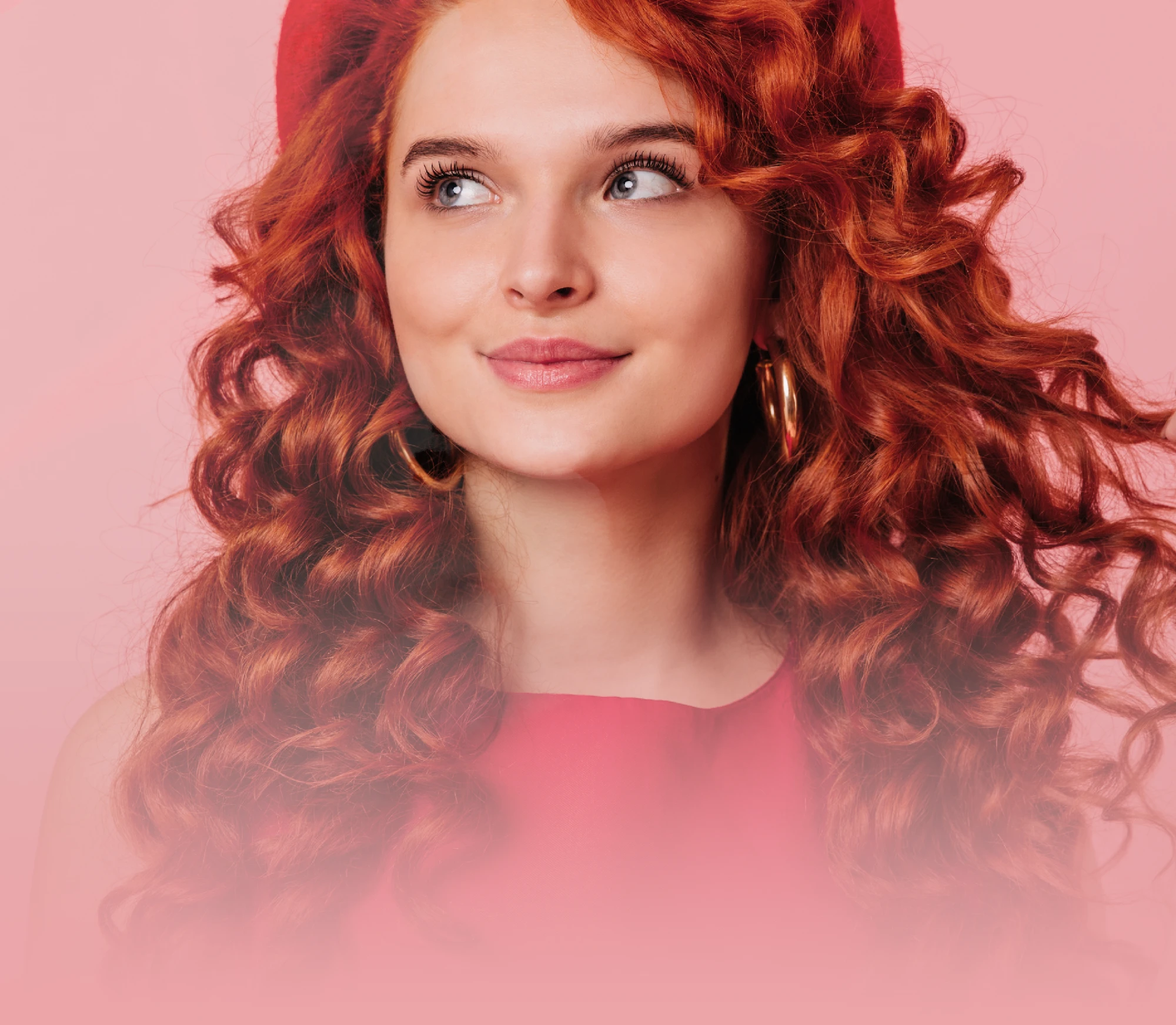 Woman with vibrant red curly hair looking to the side, wearing a red top against a soft pink studio background.