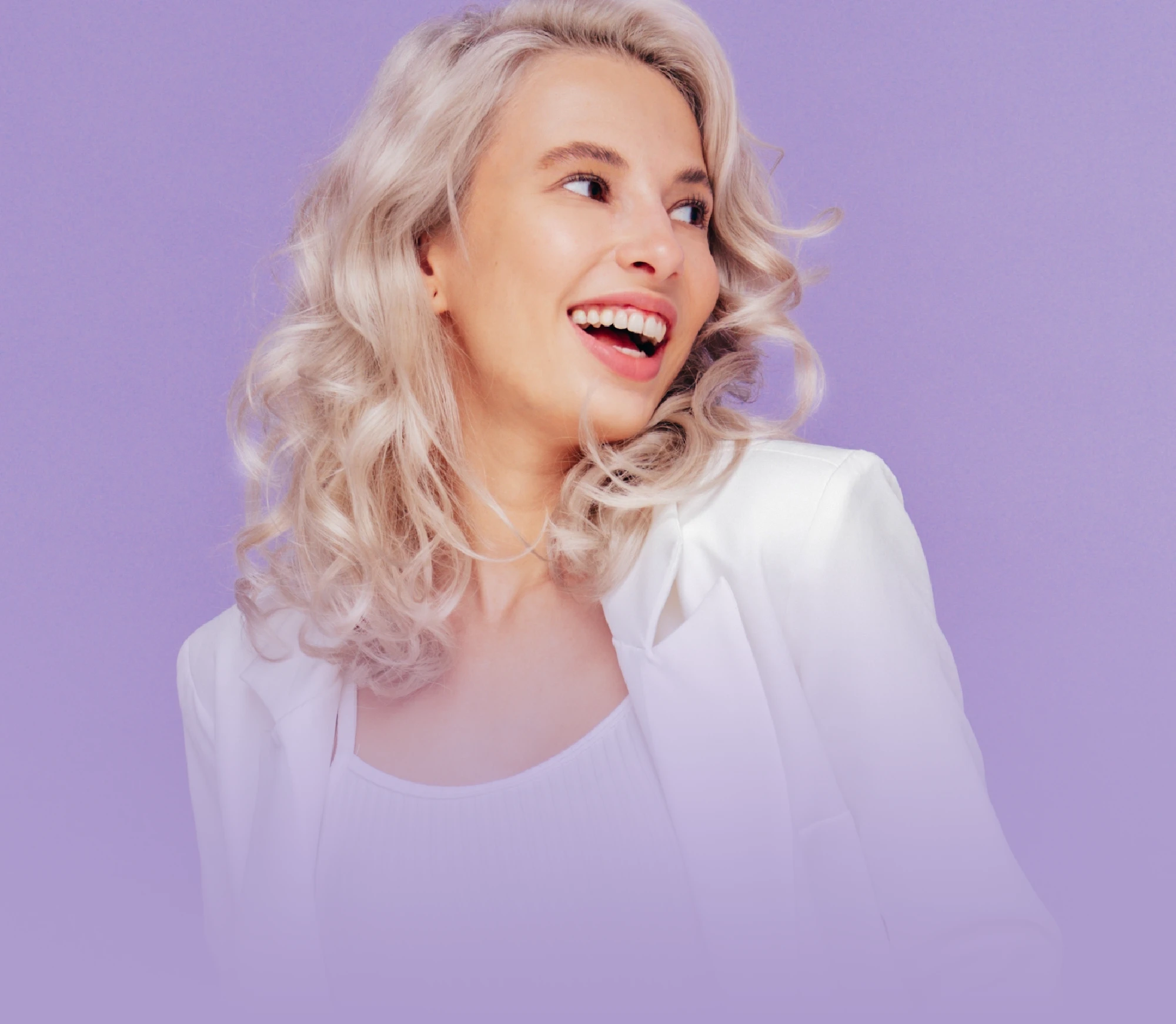 Smiling woman with silver blonde curly hair wearing a white top and jacket, looking to the side against a soft purple background.