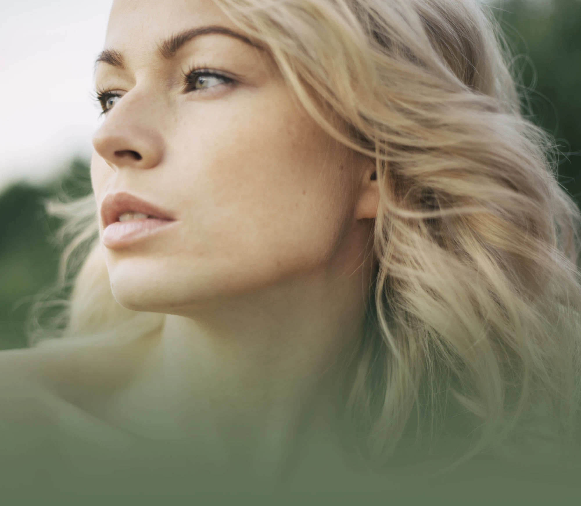 Close-up portrait of a blonde woman outdoors with soft natural light and green background tones.