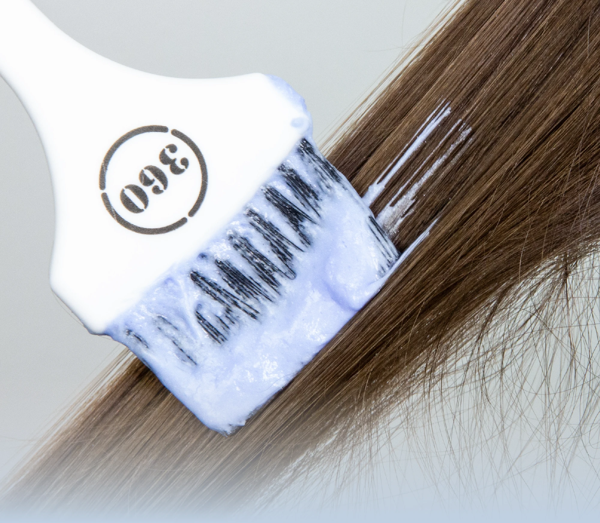 Close-up of a hair coloring brush applying light blue lightening powder cream onto a section of straight brown hair.