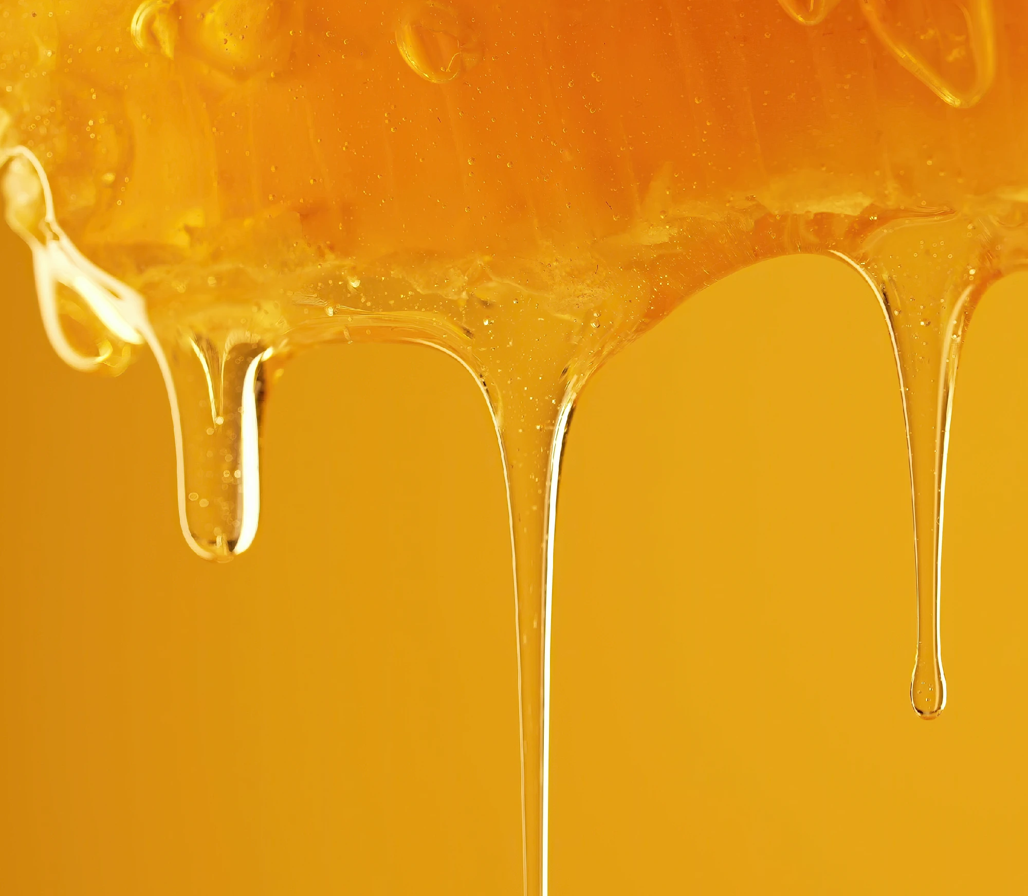 Close-up of golden honey dripping down against a warm yellow background, with thick glossy strands and droplets forming smooth vertical flows.