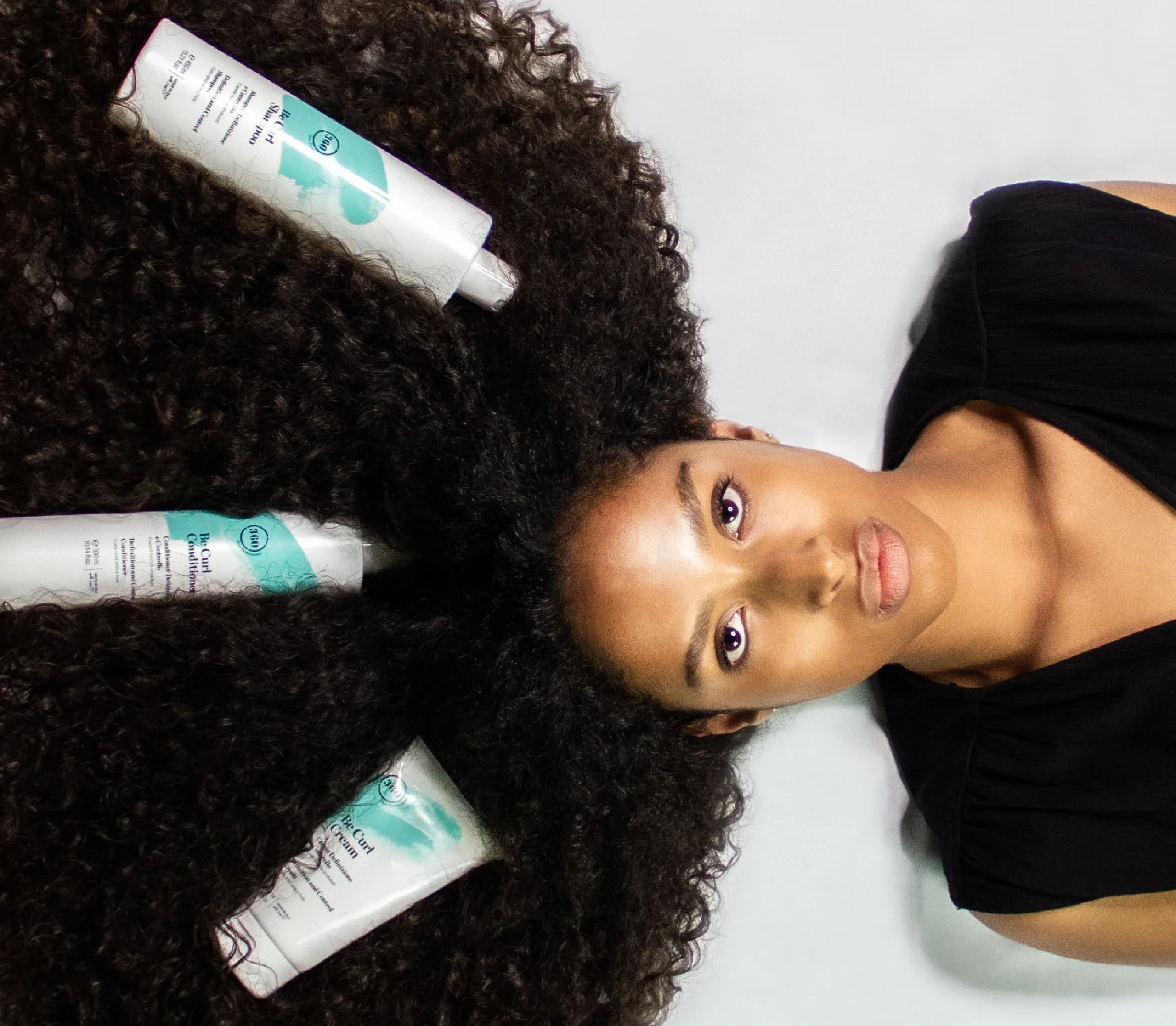 Woman with natural curly afro hair lying on a light background, surrounded by three professional haircare product bottles placed in her hair.